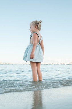 Sahilde sığ denizde duran ve bulutsuz gökyüzüne bakan, yaz elbisesi giymiş mutlu küçük bir kızın alt tarafından.