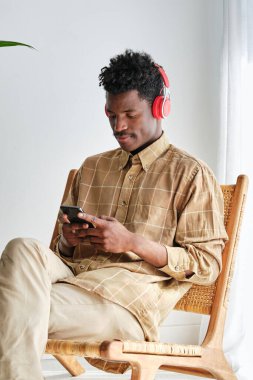 Cep telefonuyla internette sörf yaparken ve bacak bacak bacak üstüne atmış bir şekilde otururken kulaklık takan Afrikalı Amerikalı bir erkek müzik dinliyor.