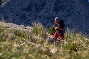 Sırt çantalı aktif bir yürüyüşçü çimenli dağ yamacında yürüyor güneşli yaz gününde doğayı keşfediyor.