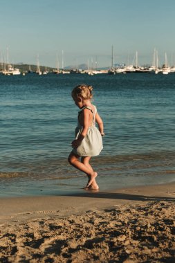 Mavi elbiseli, neşeli bir çocuğun kumlu sahilde yürüdüğünü deniz kenarında alacakaranlıkta yatlarıyla dalgalandığını görmek.