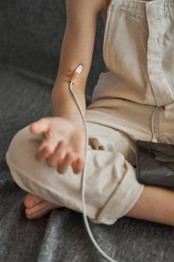 Kabloyu koluna yapışık yapıştırıcıyla şarj eden tanınamayan dijital bağımlı çocuk yerde oturuyor ve arka planda gri bir telefon var.