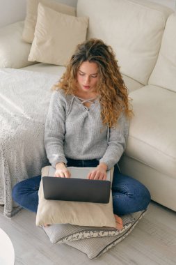 Yukardan gelen kıvırcık saçlı, sıradan elbiseli genç bir kadın yastığa oturuyor ve dizüstü bilgisayarını karıştırıyor.