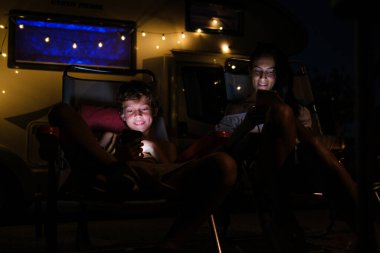Neşeli kadın ve çocuk gülümseyip cep telefonlarını karıştırırken gece karavanın dışında şezlonglarda dinleniyorlar.
