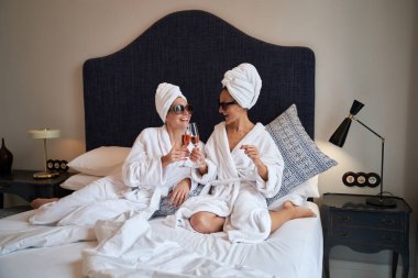 Güneş gözlüklü ve kafasında havlu türbanı olan kaplıca otelinde yatakta dinlenirken şampanya kadehleri takılan bir sürü kadın arkadaş.