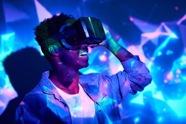 Heyecanlı Afrikalı Amerikalı erkek VR gözlük takıyor ve stüdyodaki neon ışıklarda sanal gerçekliği keşfediyor.