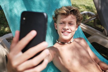 Neşeli sarı saçlı çocuk gülümseyip cep telefonunun ekranına bakarken otel bahçesinde battaniye sandalyesinde selfie çekiyor.