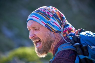 Çok renkli başörtüsü ve sırt çantası takan, sakallı orta yaşlı erkek yürüyüşçünün portresi bulanık resimli manzaranın yanında duruyor ve kapalı gözlerle bağırıyor.