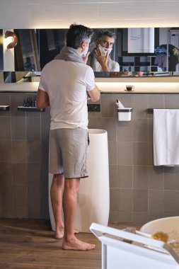 Modern banyoda ayakta duran ve sabah rutinini yaparken aynada yansımaya bakan olgun bir adam.