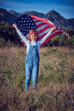 Çayırda dikilmiş kafasında Amerikan bayrağı sallayan ve kameraya bakan sıradan giyinmiş neşeli ergen çocuk.