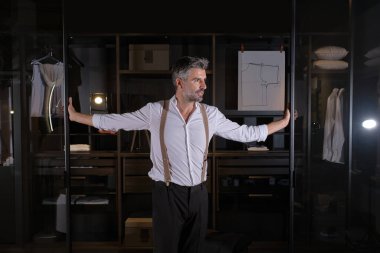 Klasik tişörtlü, beyaz sakallı adam gardırobun girişinde elleri cam sürgülü kapılar üzerinde duruyor.