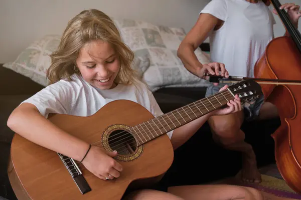 カジュアルな服を着た才能ある子供は 笑顔で ソファーの近くの床に座って 週末の自宅のリビングルームでアコースティックギターを演奏しながら下を見下ろしています — ストック写真