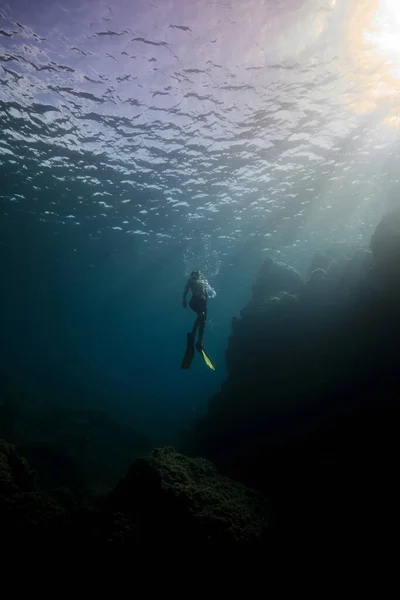 スリッパとシュノーケリングマスクのダイバーで認識できない男性ダイバーのフルボディは 石の底の近くの深い海水で泳ぎ 日光を眺めます — ストック写真