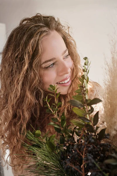 長い波状の髪を持ち 明るい部屋で緑豊かな緑の葉の近くに立って幸せそうに離れて見ている若い女性 — ストック写真