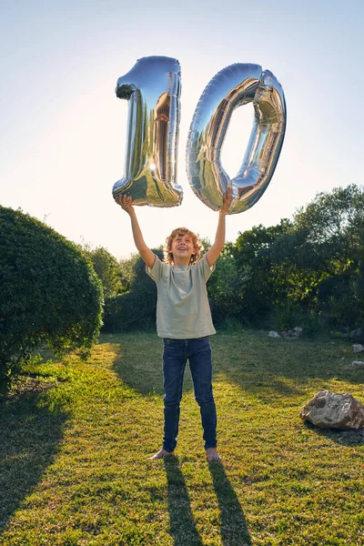 誕生日のお祝いの間に番号10の形で銀箔パーティー風船と芝生の芝生の上に立って腕を上げた笑顔の男の子の完全な体 — ストック写真