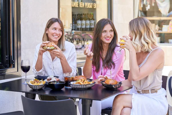 积极的女性朋友一边吃着带有薯条的开胃汉堡包 一边喝酒喝啤酒 一边聊天 相互望着对方 — 图库照片