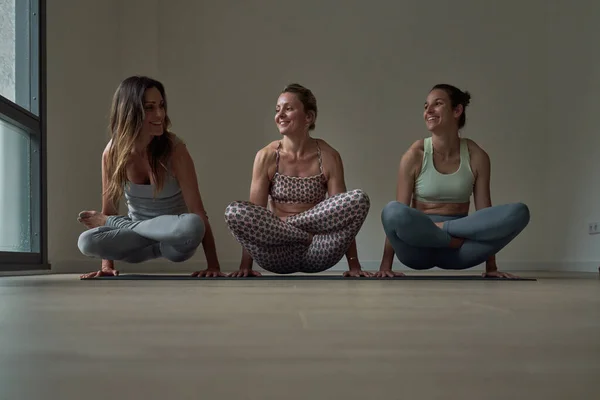 Full body of young female friends in sportswear practicing yoga and smiling while performing arm standing Tolasana pose in studio