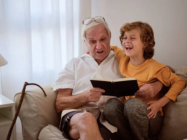 シニア男と幸せな孫座っていますソファにと面白い本を読みながら家で一緒に時間を過ごす — ストック写真