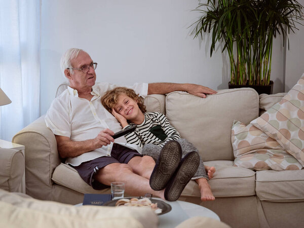 Full body of elderly man with remote control watching interesting movie with positive grandson while sitting on couch in living room