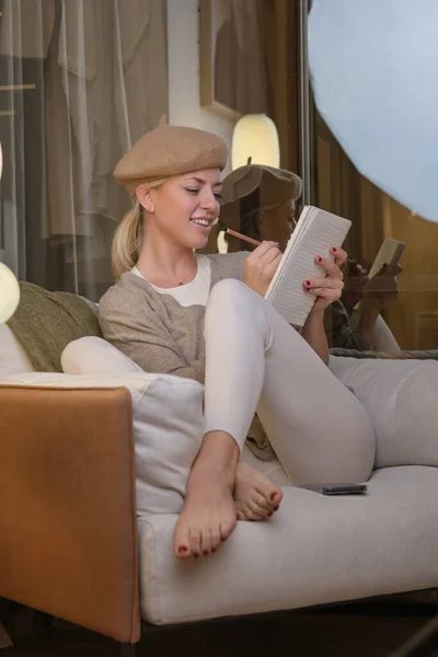 脚を交差させ ノートにスケッチを作成するアームチェアに座ってベレー帽を身に着けている陽気な若い創造的な女性アーティスト — ストック写真