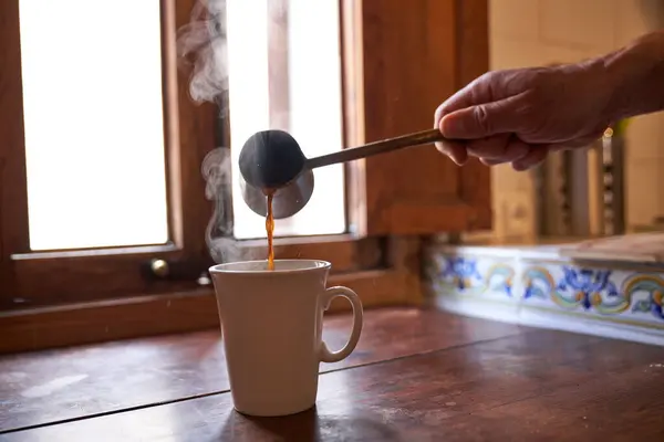 窓の近くの木製のテーブルの上に置かれた白いセラミックカップにセブから新鮮な醸造香りのコーヒーを注ぐミステリー無名の男 — ストック写真