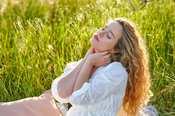 迷人的年轻女性 留着波浪般的头发 身穿白色连衣裙 轻轻地触摸着脖子 眼睛紧闭在茂密的草坪上 阳光明媚的夏日 — 图库照片