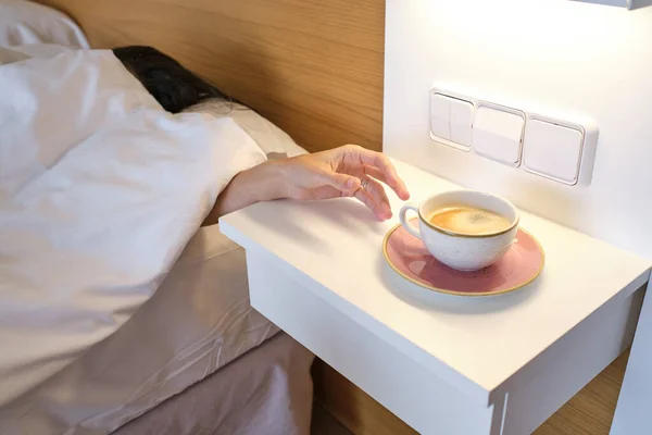 woman lying in bed and hiding under blanket while reaching out coffee mug placed on white table in bedroom at home in morning