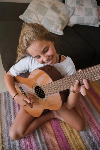 カーペットの上に座ってアコースティックギターを演奏するカジュアルな服の若いブロンドの少女の上から — ストック写真