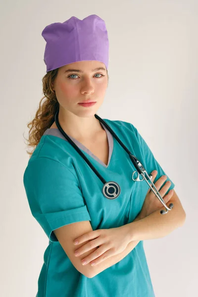 身穿军服 头戴紫色帽子 脖子上戴听诊器的年轻女子 手挽手在工作室里看着相机 — 图库照片