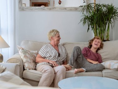 Mutlu olgun bir kadın ve gülümseyen bir çocuk kanepe ve saksı bitkisinin yanında otururken birlikte eğleniyorlar.