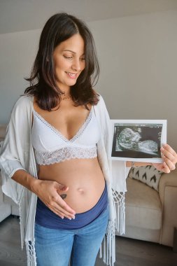 Beyaz sutyenli hamile bir kadın karnına dokunuyor ve oturma odasında dururken ultrasona bakıyor.