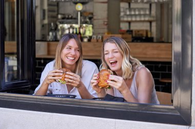 Neşeli genç kadınlar lezzetli hamburgerlerin tadını çıkarırken hafta sonu restoranda pencerenin yanında oturuyorlar.