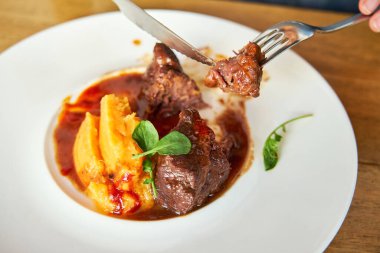 Tahta bir masadaki beyaz seramik tabakta servis edilen hindistan cevizi büyüsü ve ton balığı püresi ile kesilmiş dana yanaklarından bir parça et koparan çatalların ayrıntıları.