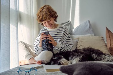 Kaliteli fotoğraf makinesi olan neşeli okul çocuğu yatakta Minyatür Schnauzer 'in yanında bacak bacak üstüne atmış oturuyor.