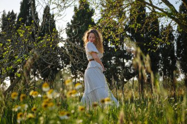 Kenar manzaralı güzel genç bir kadın Maxi yazlık elbise giyiyor güneşli kırsalda yemyeşil bir çayırda duruyor.