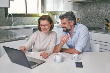 Gülümseyen yaşlı kadın, orta yaşlı sakallı bir adamla oturmuş evdeki modern mermer mutfakta bir bardak içkiyle dizüstü bilgisayarını işaret ediyor.