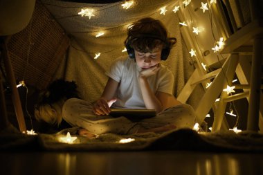 Düşük açılı, kablosuz kulaklıklı, bacak bacak üstüne atmış, parlak çelenkli, battaniyeli kaledeki tablette video izleyen ciddi bir çocuk.