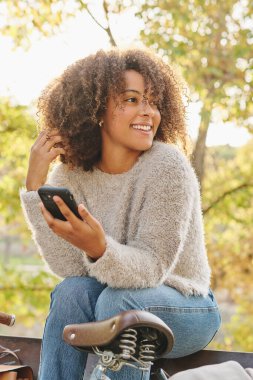 Sıcak kazaklı, kıvırcık saçlı ve kot pantolonlu pozitif İspanyol kadın bisikletinin yanında oturuyor ve interneti kolaçan ediyor.