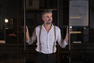 Beyaz tişörtlü, gri saçlı, orta yaşlı bir adam. Elleri cam sürgülü kapılarda dikiliyor.