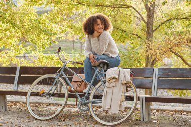 Sonbahar günü bisiklet parkının yanındaki ahşap bankta oturan, kalın kazaklı ve kot pantolonlu, pozitif, genç bir kadın vücudu.