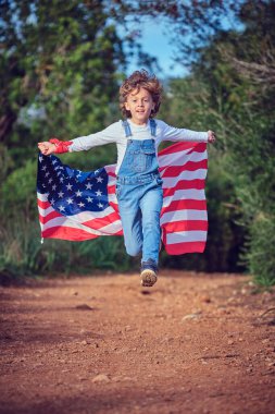 Renkli Amerikan bayrağı sallayan enerjik bir çocuk yaz günü kırsal kesimde hızla koşuyor.