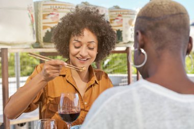 Gülümseyen Afrikalı Amerikalı kadın Japon restoranında kimliksiz siyah bir kadınla yemek yerken yemek çubuklarıyla ton balığı yiyor.