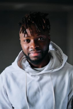 Kapüşonlu genç, gülümseyen Afro-Amerikan erkek odada karanlık arka planda dikilirken kameraya bakıyor.