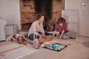 Tişörtlü, kıvırcık saçlı bir çocukla eğlenen ve kırsal evdeki kağıtta soyut resim yapan genç bir kadın sanatçı.