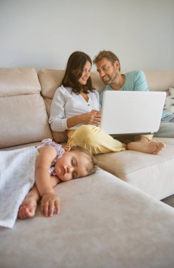 Çıplak ayaklı kadın ve erkek netbook 'ta video seyrediyor ve hafta sonu oturma odasında uyuyan kızın yanındaki koltukta oturuyor.