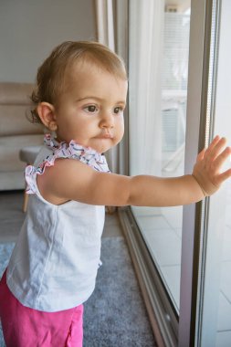 Meraklı bir çocuğun, evdeki oturma odasındaki evin etrafını incelemesi ve ilgisiyle pencereden dışarı bakmasının yan görüntüsü.