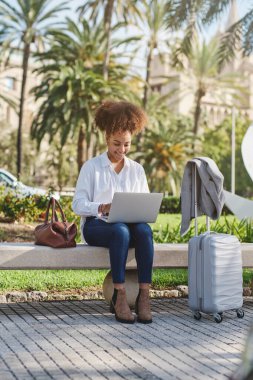 Sunny Park 'ta bankta oturan ve dizüstü bilgisayarla uzaktan kumandalı bir proje üzerinde çalışan Afrikalı Amerikalı bir kadın.