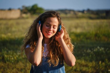 Neşeli genç bayan günlük giysiler giyiyor kulaklıkla şarkılar dinliyor ve gülümsüyor yaz parkında çimenli çimlerde kapalı gözlerle otururken