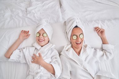 Nemlendirici salatalık dilimleri olan bornoz giymiş, cilt bakımı sırasında yatakta yatan pozitif kadın ve oğullardan.