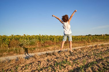Gündelik giysiler içinde, çamurlu yolda zıplayan ve ayçiçeği tarlasının yakınlarında kollarını açan kaygısız bir çocuk.