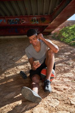 Aktif giyimli sportif İspanyol bir erkeğin tüm vücudu kumlu zeminde topuyla otururken cep telefonundan mesaj atıyor.